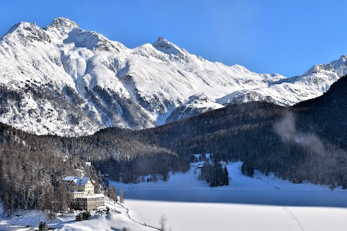 Snowy mountains in St. Moritz Switzerland, with a hotel in the foreground on the shores of a frozen-over lake.
