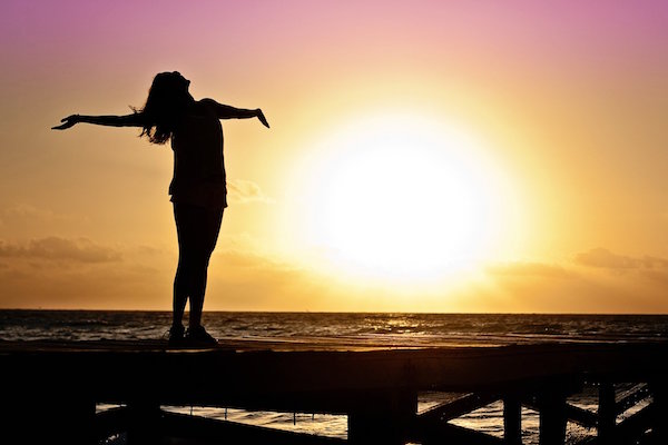 A woman looking to the sky with her arms spread wide in front of the sun rising over the ocean.