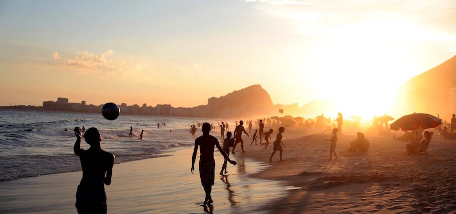 A crowded beach in Rio de Janeiro with the sun setting brightly in the background. Two boys pass a soccer ball in the foreground.